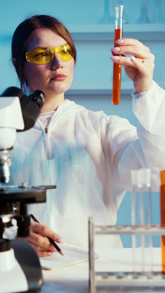 A female scientist in protective gear analyzing a test tube in a laboratory setting.